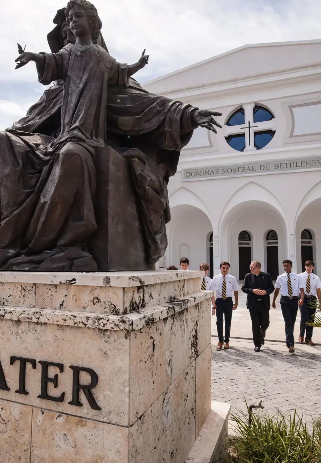 Belen Jesuit students walking in front of the Our Lady of Belen Chapel with the Alma Mater statue in the foreground.