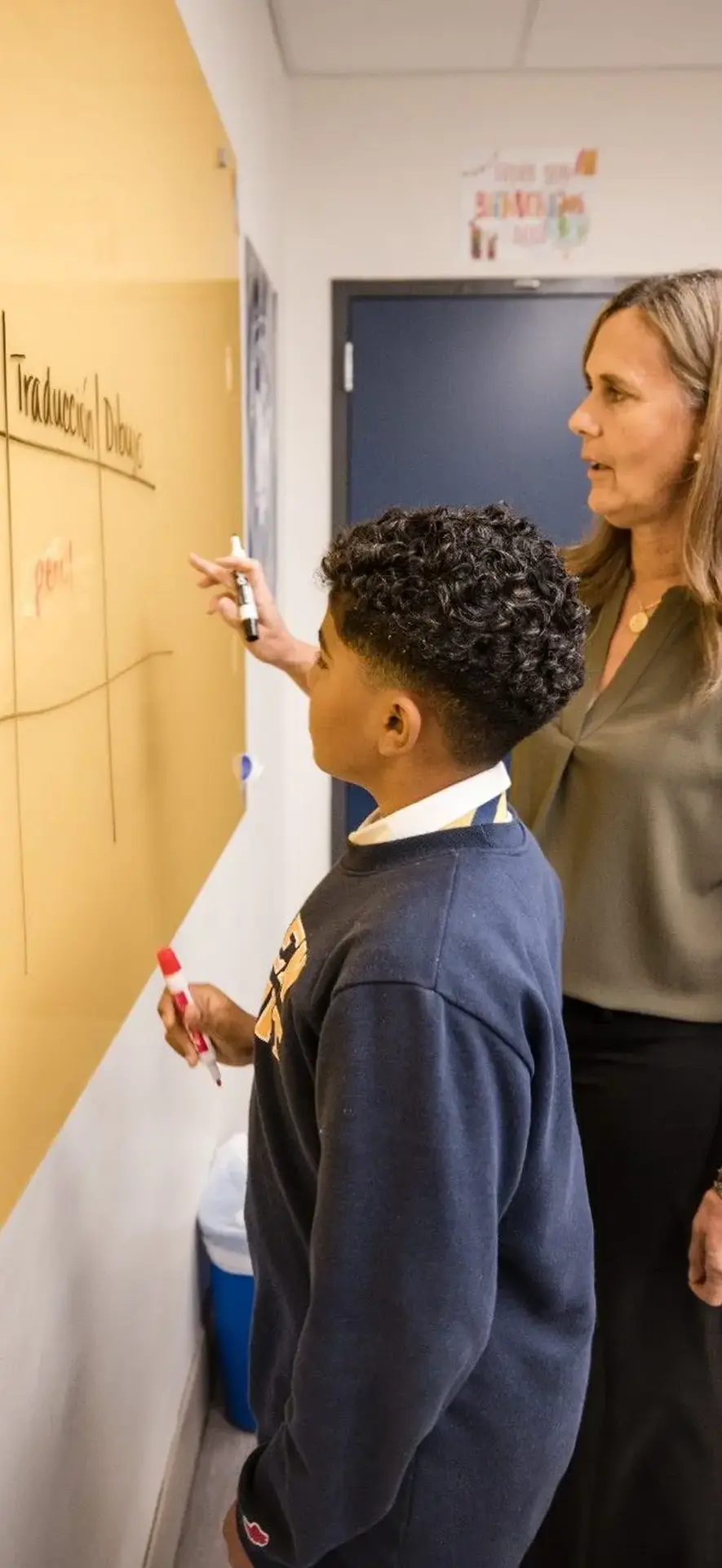 Belen Jesuit Spanish teacher Margarita Guerrero is standing beside a student, pointing to a lesson written on a large dry-erase board in the classroom.