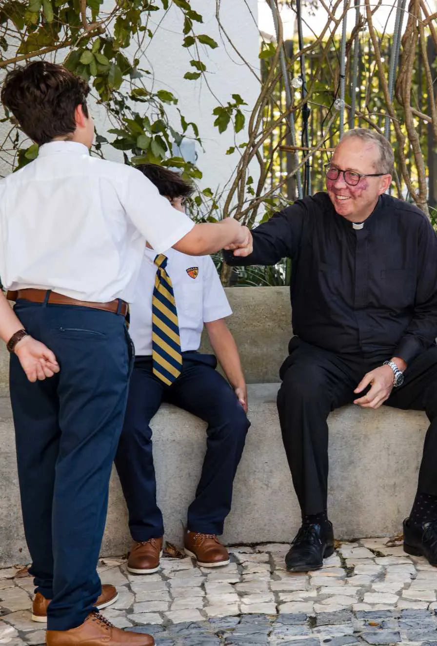 Belen Jesuit President Fr. Willie Garcia-Tunon, S.J., is seated, smiling, and fist-bumping a middle school student while engaging in conversation with four students.