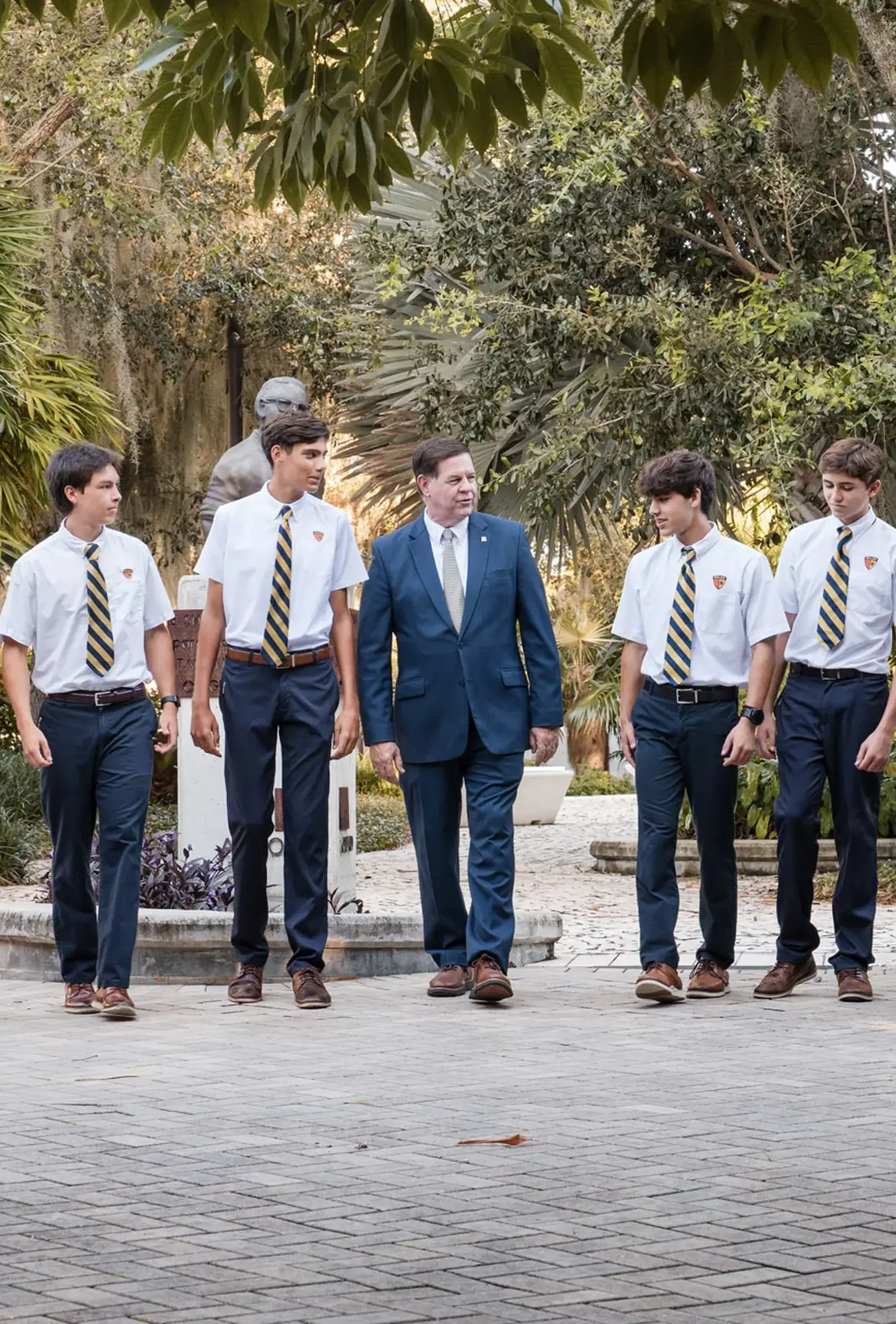 Belen Jesuit Principal Jose E. Roca walks and converses with six high school students in the Arroyo Quad on a sunny day.