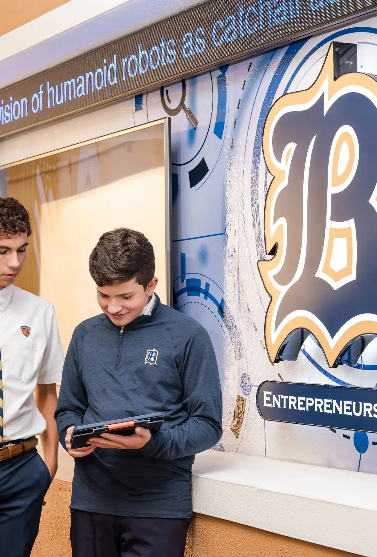 Two Belen Jesuit students stand outside the Catholic Entrepreneurship Program classroom, reviewing notes on an iPad. Visible are a stock market ticker, large screens displaying business news, and the 